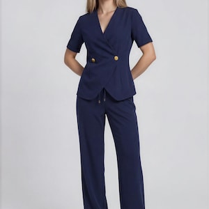May include: Navy blue two-piece suit. The short-sleeved blazer features a wrap-style front with two gold buttons. The matching pants are wide-legged. The outfit is paired with white sneakers. A modern and stylish ensemble.