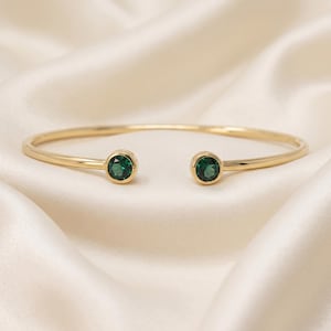 Può includere: Un bracciale aperto color oro con due pietre rotonde sfaccettate verde scuro, incastonate in castoni di metallo lucido su ciascuna estremità. Il bracciale presenta una fascia sottile e liscia che si curva in forma circolare, mettendo in risalto le pietre.
