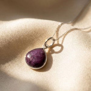 Può includere: Una collana d'argento con un ciondolo a forma di goccia. Il ciondolo contiene una pietra preziosa viola scuro, incastonata in una lunetta d'argento, sospesa a una sottile catena d'argento. La pietra ha una finitura lucida.