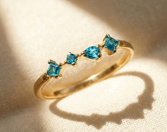 May include: A gold ring featuring four blue gemstones in square and teardrop shapes. The gemstones are set along the top of the ring band. The ring is displayed on a white surface.