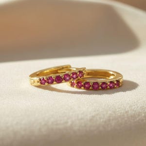 Puede incluir: Dos pendientes de aro dorados con una fila de pequeñas piedras preciosas de color rosa brillante. Los pendientes se presentan sobre un fondo blanco roto, resaltando su color y diseño. Joyería clásica.