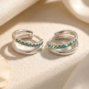 May include: A pair of silver-toned hoop earrings featuring a double-band design. The outer band of each hoop is embellished with a row of small, round, vibrant green gemstones, while the inner band remains a plain, polished silver-toned metal.