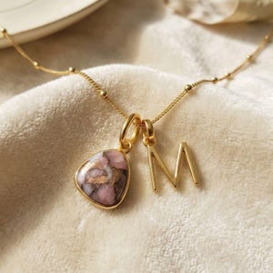 May include: A gold necklace with a pink and gold stone pendant and a gold letter "M" pendant. The chain has small gold beads. The pendants hang on a soft, off-white surface. The necklace is a delicate piece of jewellery.