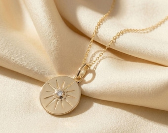 May include: A gold-colored pendant necklace featuring a circular pendant with a sunburst design and a small, clear gemstone at the center. The necklace chain is delicate and gold, resting on a cream-colored fabric.