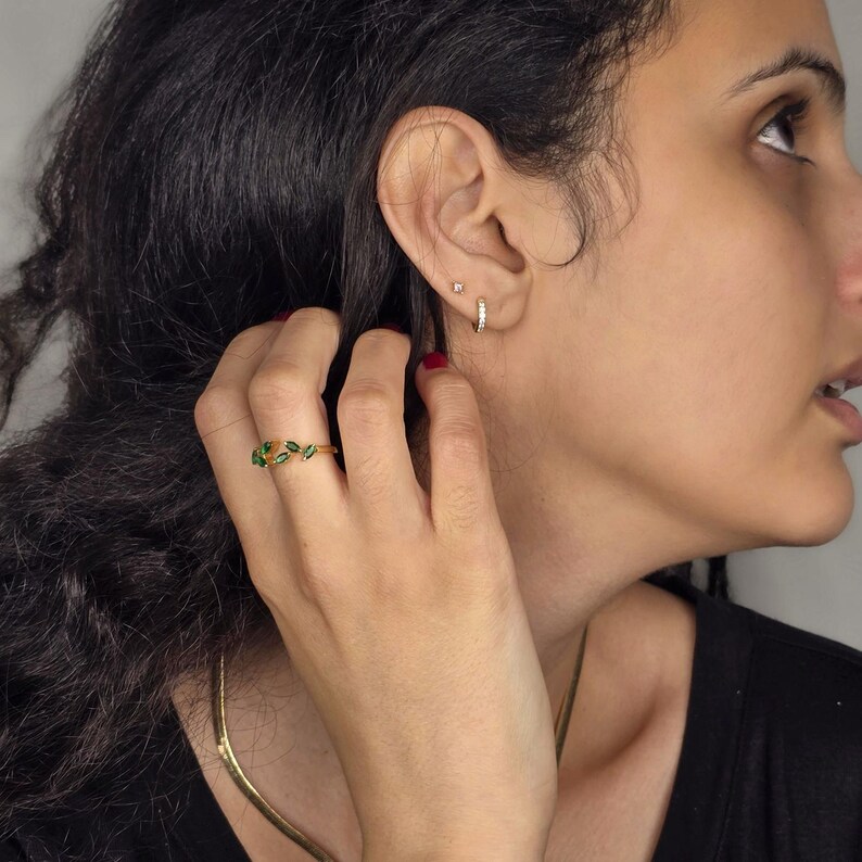 May include: A gold ring with green leaf accents. The ring is on a person's finger. The person is also wearing a gold necklace and small gold hoop earrings. The person is wearing a black top.
