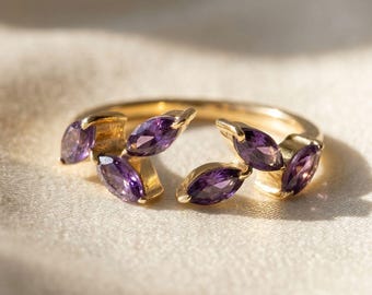 May include: A gold ring featuring marquise-cut purple gemstones. The ring's design incorporates the gemstones in a leaf-like arrangement. The ring is set against a soft, off-white background, highlighting the gold and purple hues.