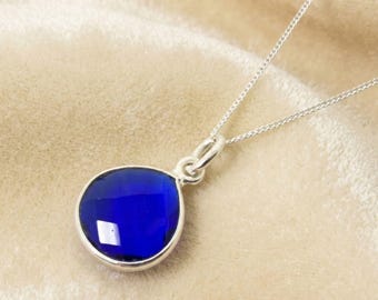 Könnte beinhalten: Eine silberne Halskette mit einem auffälligen, tiefblauen Anhänger. Der Anhänger hat eine abgerundete Form und eine facettierte Oberfläche. Die Kette ist filigran und der Anhänger ist in eine silberne Lünette gefasst. Die Farbe des Anhängers ist ein sattes Saphirblau.