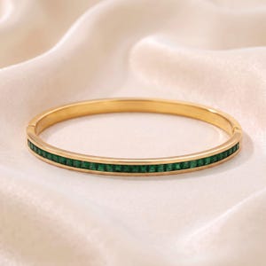 Könnte beinhalten: Ein goldfarbener Klapparmreif mit einer durchgehenden Reihe kleiner, quadratisch geschliffener smaragdgrüner Steine, die in die Mitte des Bandes eingelassen sind. Das Armband hat eine polierte Metalloberfläche und einen sicheren Verschluss.
