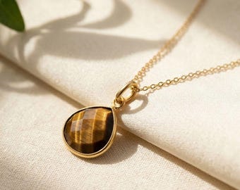 May include: A gold-colored necklace with a teardrop-shaped pendant. The pendant features a faceted, brown tiger's eye stone set in a gold bezel. The chain is delicate and the background is a soft, off-white fabric.