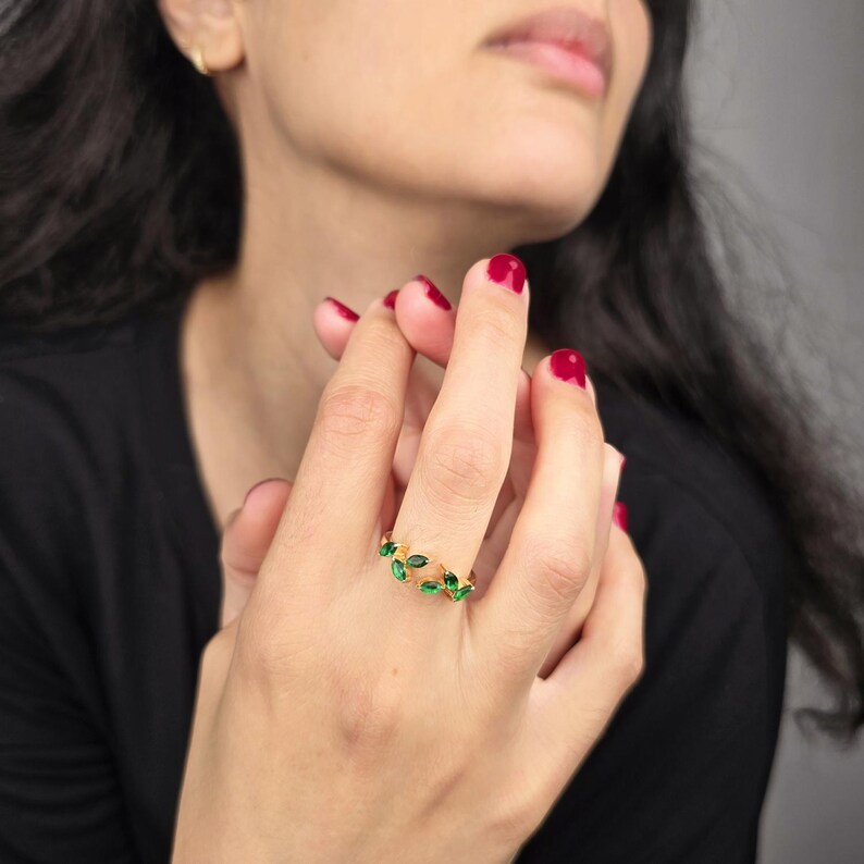 May include: A gold-colored ring featuring a leaf design with green gemstones. The ring is worn on a finger, showcasing the intricate details of the jewelry. The gemstones are emerald green, adding a pop of color to the gold band. The ring is a piece of fashion jewelry.