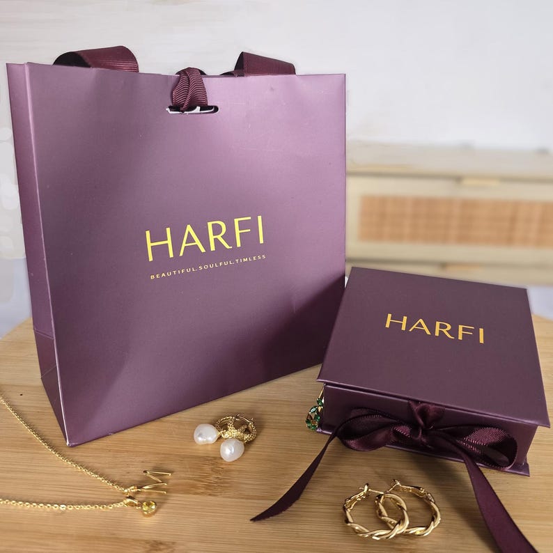 May include: A purple gift bag and box with the gold text "HARFI" and "BEAUTIFUL SOULFUL TIMELESS". Gold jewelry, including a necklace, earrings, and hoop earrings, are displayed on a wooden surface.