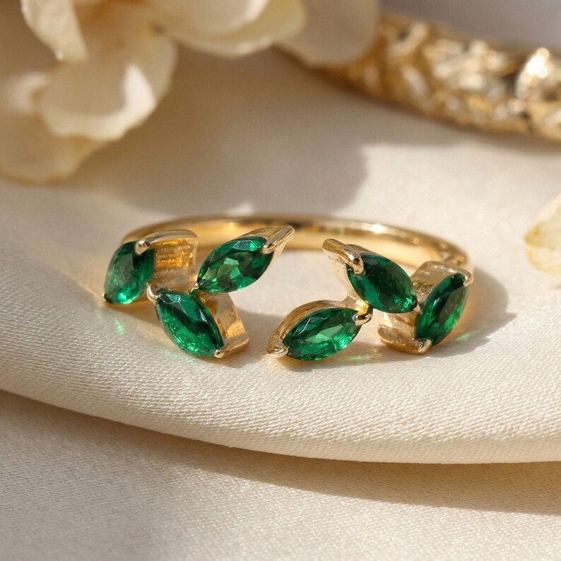 May include: A gold ring featuring a leaf design with marquise-cut emerald green gemstones. The ring is set on a white surface, with a soft focus background of white and gold elements.