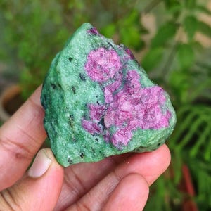 Puede incluir: Una gema en bruto con una vibrante combinación de colores. La piedra presenta una base verde moteada con parches magenta intenso. Pequeñas inclusiones negras salpican la superficie, agregando textura a este espécimen mineral natural.