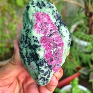 Peut inclure: Une pierre précieuse brute avec un centre rose vif entouré de zones vertes et noires mouchetées. La pierre naturelle est tenue dans une main, mettant en valeur sa texture et ses variations de couleurs uniques. Le minéral est naturel.