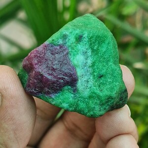 Könnte beinhalten: Ein roher Edelstein, überwiegend grün mit einem auffälligen Fleck in tiefem Rot-Violett. Der Stein wird in einer Hand gehalten, mit einem verschwommenen grünen Hintergrund.