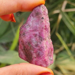 May include: A raw, uncut ruby gemstone. The ruby is a deep pinkish-red colour with darker inclusions, held between fingers. The natural stone has a textured surface and irregular shape, showcasing its raw, unpolished state.