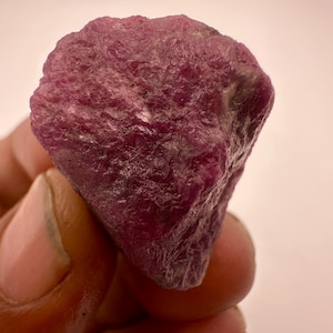 May include: A raw, unpolished ruby gemstone with a deep magenta and purple hue. The stone features a naturally textured, uneven surface with crystalline facets and opaque sections, showcasing its organic, uncut form and irregular shape.