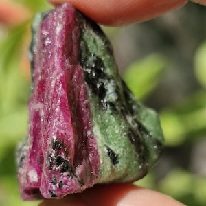 May include: A close-up of a rough ruby crystal, held between fingers. The stone displays a vibrant mix of deep red and green hues, with black speckles. The natural gemstone has a raw, unpolished texture, showcasing its mineral composition.