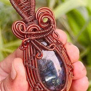 May include: A copper wire wrapped pendant featuring a dark grey and green agate stone. The pendant has a unique, intricate design with a heart-shaped top.