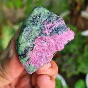 May include: A close-up of a rough gemstone with a mix of colours. The stone features areas of vibrant pink, deep green, and black, creating a striking contrast. The texture appears rough and natural, showcasing the stone's raw beauty.