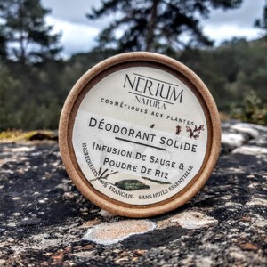 Peut inclure: Un contenant de déodorant solide rond avec l'inscription "NERIUM NATURA" et "DÉODORANT SOLIDE". L'étiquette indique également "INFUSION DE SAUGE & POUDRE DE RIZ". Le contenant est en carton et repose sur une surface rocheuse texturée.