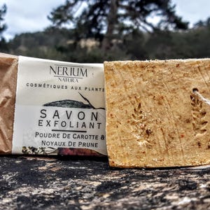 Può includere: Una saponetta esfoliante e una saponetta avvolta. La saponetta avvolta ha un'etichetta che recita "NERIUM NATURA COSMÉTIQUES AUX PLANTES SAVON EXFOLIANT POUDRE DE CAROTTE & NOYAUX DE PRUNE". La saponetta non avvolta è marrone chiaro con un disegno di grano.