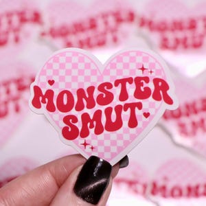 K&ouml;nnte beinhalten: Ein herzf&ouml;rmiger Aufkleber mit dem Text "MONSTER SMUT" in roter, retroartiger Schrift. Das Herz hat ein rosa-wei&szlig;es Karomuster. Weitere Aufkleber mit dem gleichen Design befinden sich im Hintergrund.