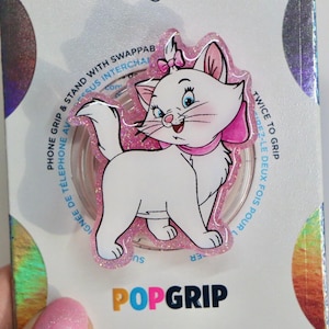 May include: A pink and white phone grip with a glittery background featuring a cartoon image of a white cat with pink ears and a pink bow. The text "POPGRIP" is printed in pink, blue, and yellow letters below the image.