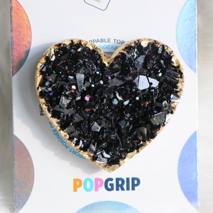 May include: Black heart-shaped phone grip with gold trim and a glittery finish. The grip is made of resin and has a textured surface. The grip is designed to be used with a smartphone or tablet. The grip is also compatible with most cases. The grip is available in a variety of colours and designs. POPGRIP.