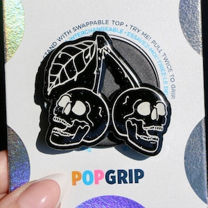 Peut inclure: Poignée de téléphone en forme de cerise avec des crânes noirs et blancs avec le texte "STAND WITH SWAPPABLE TOP. TRY ME! PULL TWICE TO GRIP. INTERCHANGEABLE. ESSAYEZ-MOITTIREZ-LE DE" et le logo "POPGRIP".