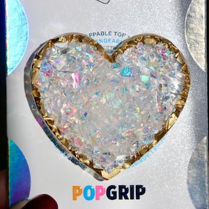 May include: A gold heart-shaped phone grip with iridescent glitter embedded inside. The grip is labeled "POPGRIP" in colorful letters.