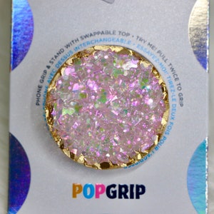 May include: A pink and gold phone grip with a glittery, iridescent finish. The grip is round and has a gold rim. The text "POPGRIP" is printed on the packaging below the grip.