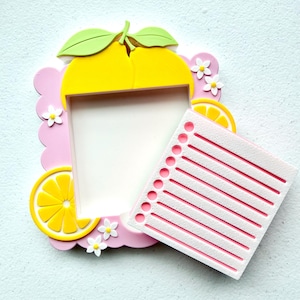 May include: A decorative pink picture frame with a lemon theme. The frame has a scalloped edge, yellow lemons, green leaves, and small white flowers. A notepad-shaped piece with pink lines is included.