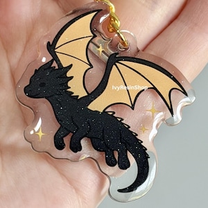 May include: A black glitter dragon keychain with tan wings and gold accents. The dragon is a cartoon style design with a clear background and gold-colored hardware. The keychain is a fantasy-themed accessory.