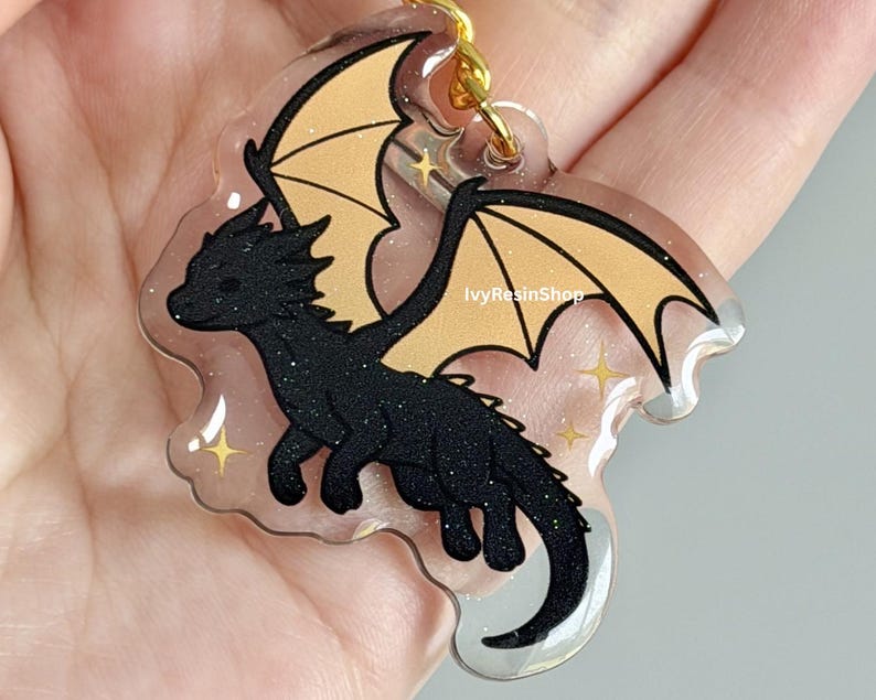 May include: A black glitter dragon keychain with tan wings and gold accents. The dragon is a cartoon style design with a clear background and gold-colored hardware. The keychain is a fantasy-themed accessory.