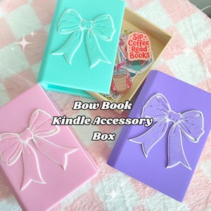May include: A collection of Bow Book Kindle Accessory Boxes in pastel colors: teal, pink, and purple. Each box features a 3D bow design on the cover. The boxes are displayed on a pink and white checkered surface.
