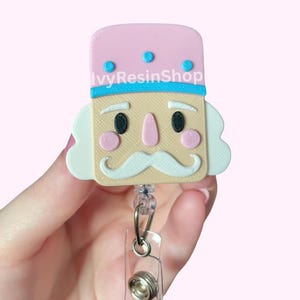 May include: A pink and beige badge reel featuring a cartoon nutcracker design. The nutcracker has a pink hat with blue accents, a beige face, and a white mustache. The text "Ivy Resin Shop" is visible on the top of the badge reel.
