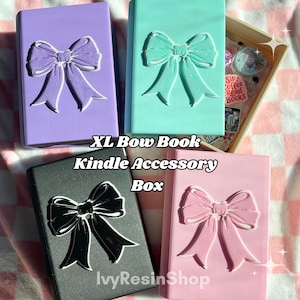 May include: Four Kindle accessory boxes in pastel colors: lavender, mint, black, and pink. Each box features a large bow design. The text "XL Bow Book Kindle Accessory Box" is visible. A wooden box with stickers is also present.