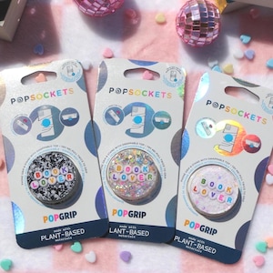 Peut inclure: Trois supports de téléphone PopSockets avec le texte "BOOK LOVER". Les designs incluent du noir et blanc, des paillettes et des couleurs pastel. Chaque support est emballé sur une carte blanche et bleue avec le logo PopSockets.