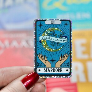 Pu&ograve; includere: Un adesivo olografico con sfondo blu e una luna crescente con un design di glitter dorati. L'adesivo ha il testo "light it up!" su uno striscione e "STARBORN" in basso. L'adesivo &egrave; una carta dei tarocchi con il numero XII.