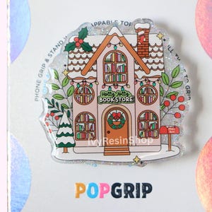 May include: A PopGrip phone grip with a Christmas bookstore design. The image shows a snow-covered roof, a wreath, and the text "Holly Jolly Bookstore." The grip is white with glitter accents and a festive theme.