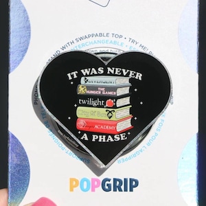 以下が含まれることがあります： 「IT WAS NEVER A PHASE」の文字と、Divergent、The Hunger Games、Twilight、City of Bones、Academyの書籍タイトルが書かれた、黒いハート型のPopGrip。PopGripは白と虹色の背景にあります。
