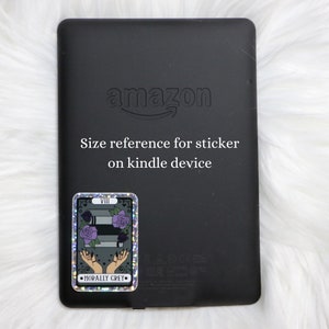 May include: A black Amazon Kindle e-reader with a holographic sticker on the back. The sticker features a tarot card with the number VIII, a stack of books, and the text "Morally Grey".