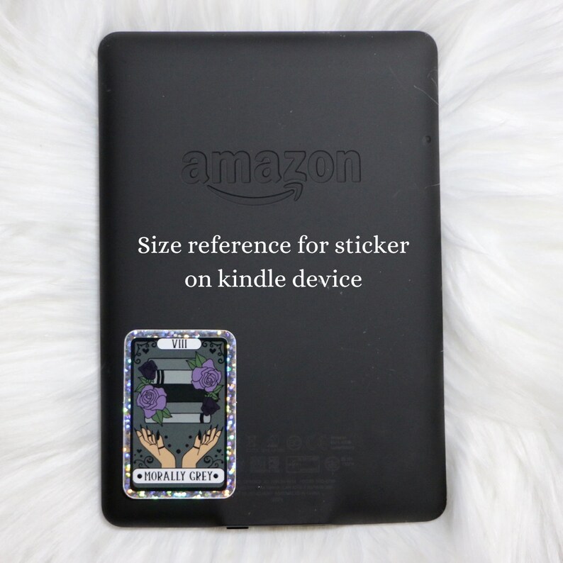 May include: A black Amazon Kindle e-reader with a holographic sticker on the back. The sticker features a tarot card with the number VIII, a stack of books, and the text "Morally Grey".