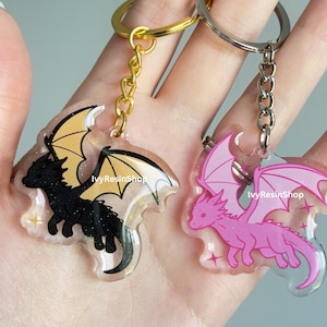 May include: Two dragon-shaped keychains. One is black with gold wings, the other is pink with pink wings. Both have a clear background with star accents and gold or silver key rings. The text "IvyResinShop" is visible.