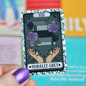 May include: A tarot card with a glittery silver border and a black and white illustration of a stack of books with purple flowers and two hands with black nails. The card is labeled "VIII" and "MORALLY GREY".