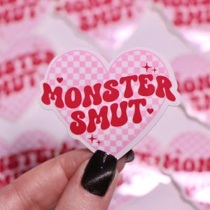 K&ouml;nnte beinhalten: Ein pinkfarbener herzf&ouml;rmiger Aufkleber mit einem Karomuster und den Worten "MONSTER SMUT" in roter Schrift im Retro-Stil. Der Aufkleber hat einen wei&szlig;en Rand und wird vor anderen &auml;hnlichen Aufklebern gehalten.