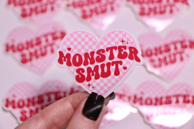K&ouml;nnte beinhalten: Ein pinkfarbener herzf&ouml;rmiger Aufkleber mit einem Karomuster und den Worten "MONSTER SMUT" in roter Schrift im Retro-Stil. Der Aufkleber hat einen wei&szlig;en Rand und wird vor anderen &auml;hnlichen Aufklebern gehalten.