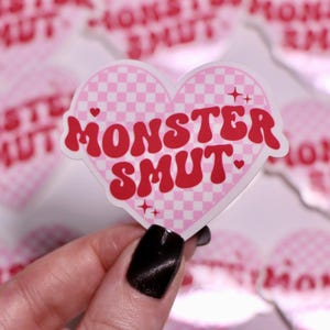 K&ouml;nnte beinhalten: Ein herzf&ouml;rmiger Aufkleber mit einem rosa-wei&szlig; karierten Muster und dem Schriftzug "MONSTER SMUT" in roter, retroartiger Schrift. Der Aufkleber hat kleine Stern- und Herzakzente.
