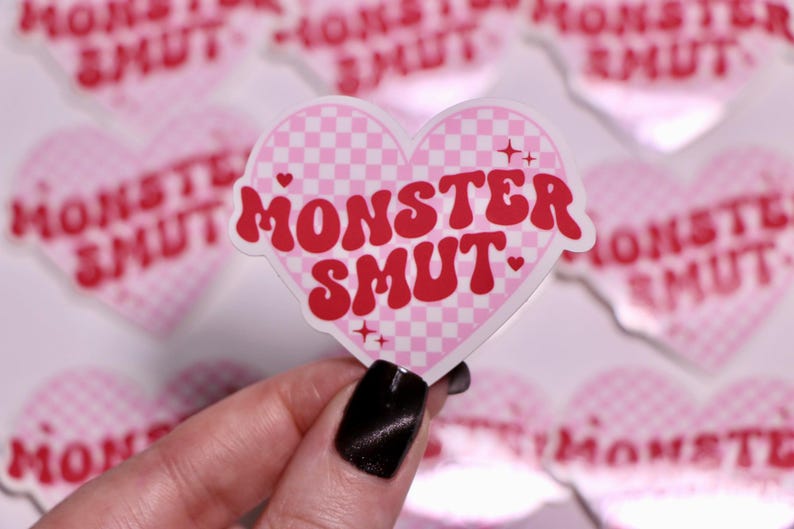 K&ouml;nnte beinhalten: Ein herzf&ouml;rmiger Aufkleber mit einem rosa-wei&szlig; karierten Muster und dem Schriftzug "MONSTER SMUT" in roter, retroartiger Schrift. Der Aufkleber hat kleine Stern- und Herzakzente.